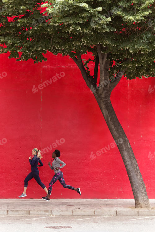 Preview: Two sporty young women running together in the city passing red wall