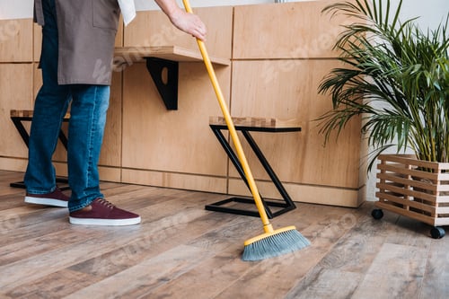 Preview: partial view of barista in apron cleaning floor with broom in cafe