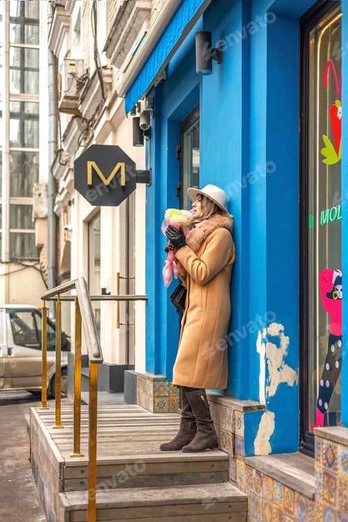 Preview: A woman standing alone against a wall near a store with a bouquet. She wears a brown coat and hat