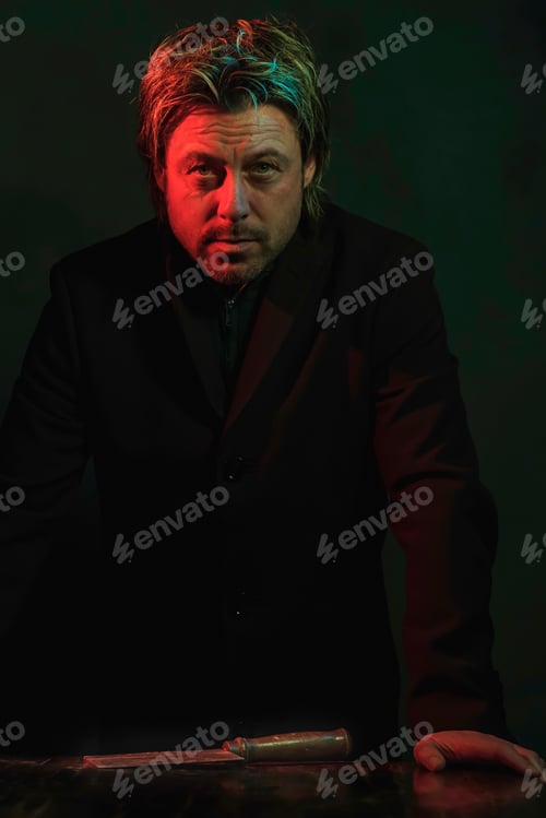 Preview: Man Standing At Table With Meat Cleaver On It. Illuminated By Red And Green Light.