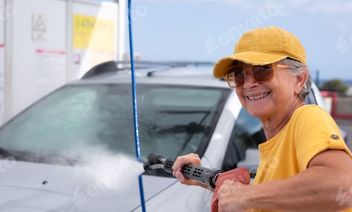 Preview: Cheerful active senior woman washing her car in a self-service car wash station using high pressure