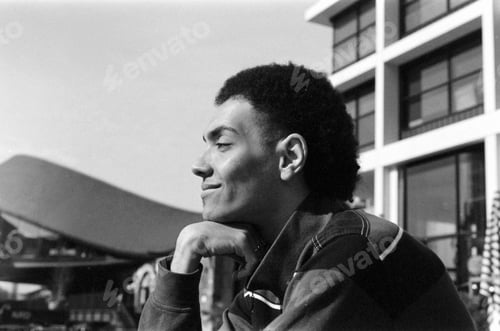 Preview: Young man smiling and resting chin on hand in urban setting, grainy analog photo