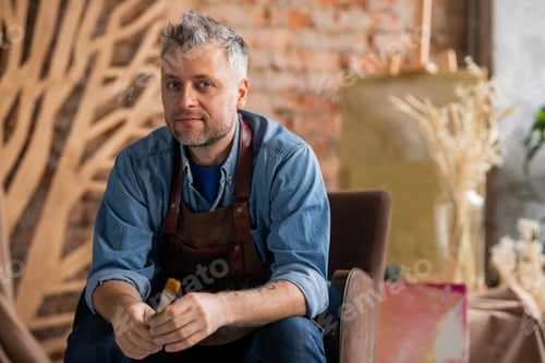 Preview: Successful creative painter with paintbrush sitting in armchair in modern studio