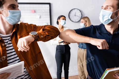 Preview: young managers in medical mask doing elbow bump gesture near businesswomen on blurred background