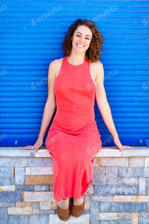 Preview: Happy woman in red dress sitting on stone wall