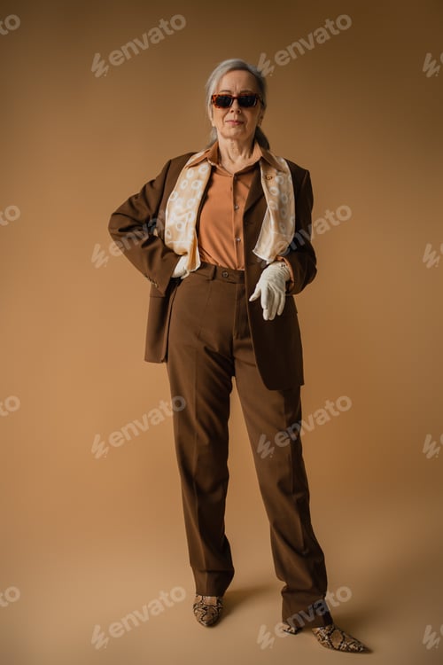 Preview: full length of senior woman in brown suit and white gloves posing with hand on hip on beige