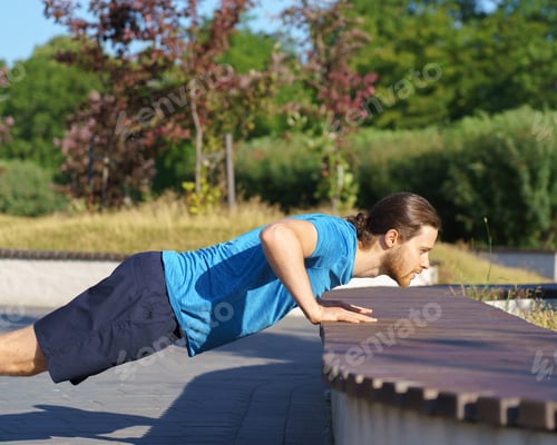 Preview: Sportive guy training arms while doing push ups on bench in park on summer day