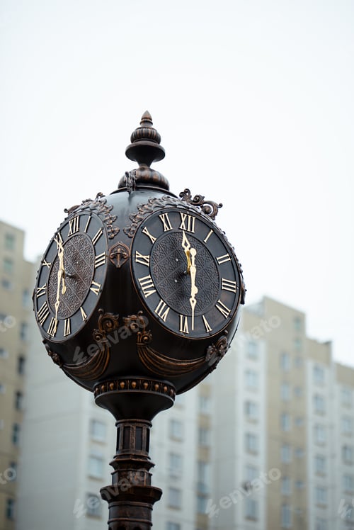 Preview: The dial of a street metal brown clock with Roman numerals in close-up on the background of a house.