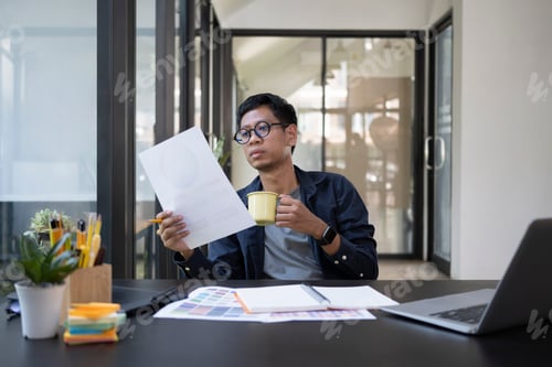 Preview: Young man graphic designer drinking coffee and working with color swatch in creative office.
