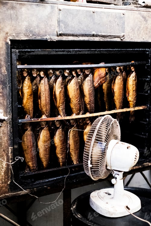 Preview: Close up of rows of freshly smoked whole trout in a smoker, white fan standing in front.