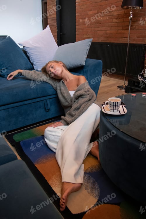 Preview: fashionably dressed blonde girl sits on the floor near the sofa in the living room and is sad.