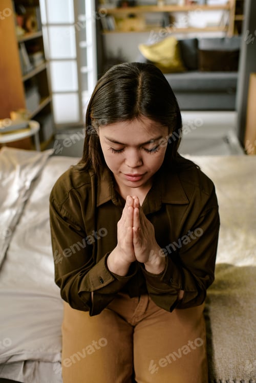 Preview: Young Adult Asian Woman Sitting on Bed Praying with Eyes Closed Indoors