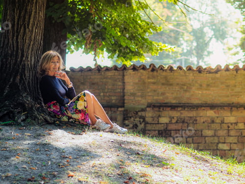 Preview: senior fit happy lady relax sit next to tree in park wear floral skirt and black long sleeve shirt
