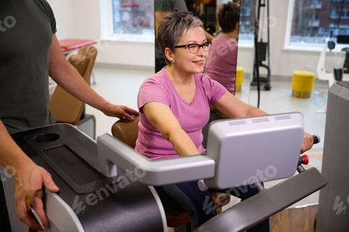 Preview: Trainer helping adult woman doing exercise for legs on sport equipment in gym