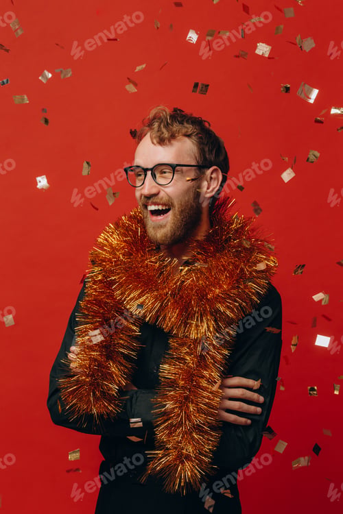 Preview: Red-haired cheerful man in glasses and golden boa at New Year party with confetti. Studio photo.