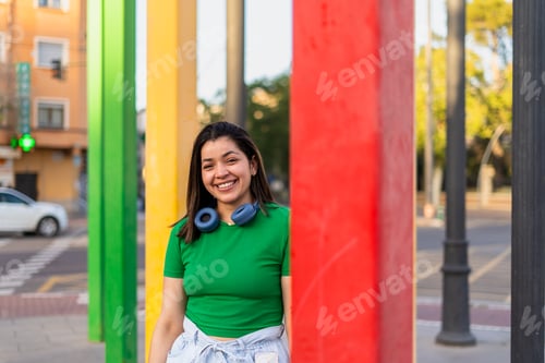 Preview: Young woman smiling with headphones around neck by colorful poles in the city