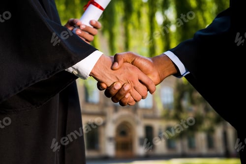 Preview: Congratulations! Close-up of two men in graduation gowns holding diplomas and shaking hands