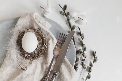 Preview: Stylish Easter table setting flat lay. Easter egg in nest, willow branches, feathers on modern plate