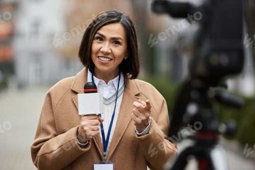 Preview: Multiracial journalist in beige coat reporting live with microphone in an urban setting