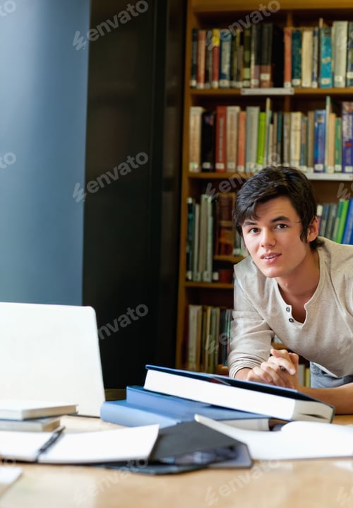 Preview: Male student in his 20s leaning over table, reading textbooks with laptop and notes, copy space