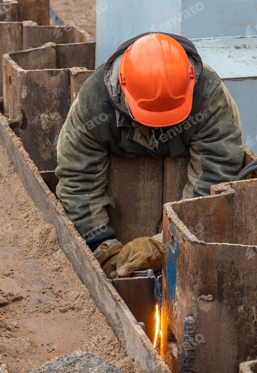 Preview: a builder in an orange helmet is engaged in welding on a construction site, strengthening the metal