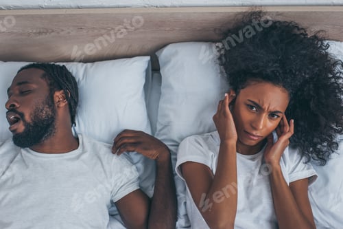 Preview: top view of african american woman suffering from headache while lying in bed near snoring husband