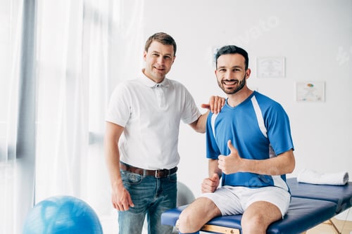 Preview: smiling Physiotherapist near happy football player showing thumb up in hospital