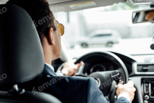 Preview: back view of african american businessman in suit and sunglasses driving car