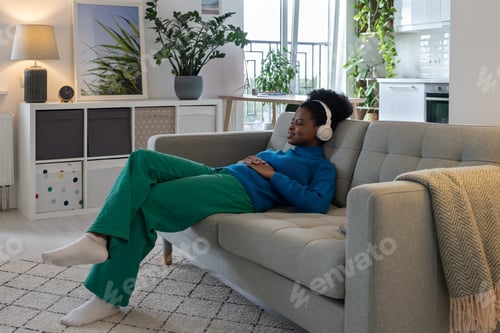 Preview: Happy relaxed African American woman in headphones closing eyes enjoying music sits on sofa