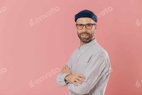 Preview: Studio shot of self assured bearded man with blue eyes, wears stylish hat and white shirt