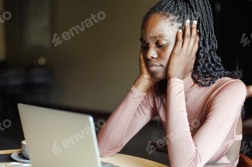 Preview: Portrait of young tired African-American woman sitting at table, looking at screen of laptop