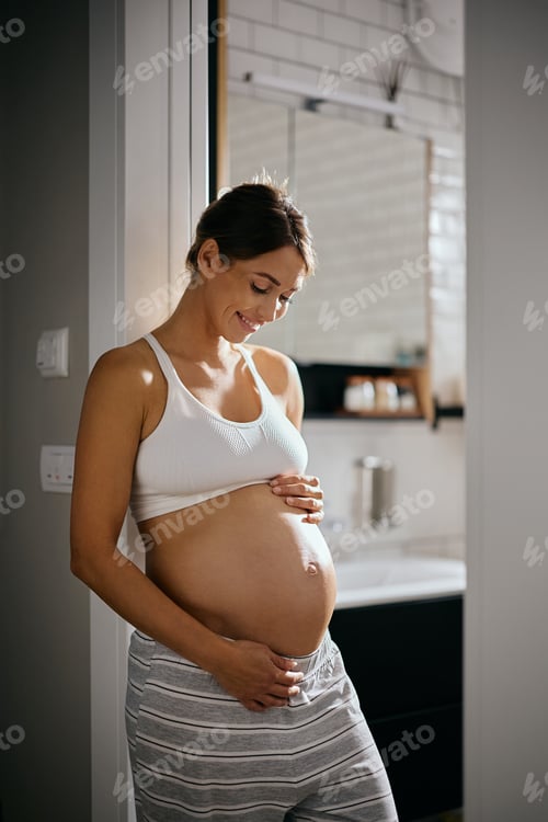 Vista previa: Feliz mujer embarazada mirando su estómago en casa.
