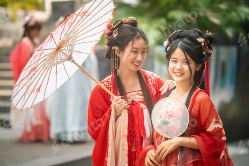 Preview: Celebration of traditional culture with two women dressed in red silk attire in a garden setting