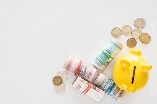 Preview: elevated view of yellow piggy bank and various rolled russian banknotes with coins on white surface