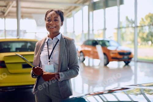 Preview: Happy black saleswoman at car dealership looking at camera.