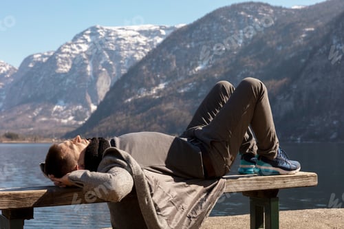 Preview: Young man lying outdoors on bench and enjoying mountains, snow, good weather, blue sky, sun.