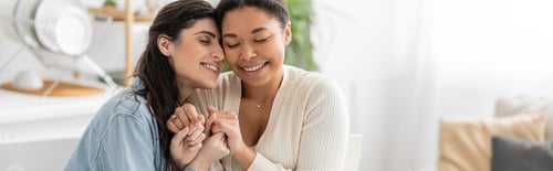 Preview: pleased interracial lesbian couple with closed eyes holding hands at home, banner