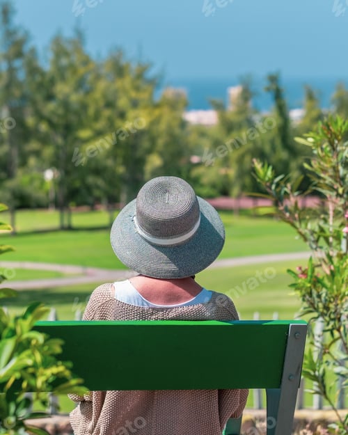 Preview: Rear view of elderly caucasian woman with hat sitting on outdoor bench in public park