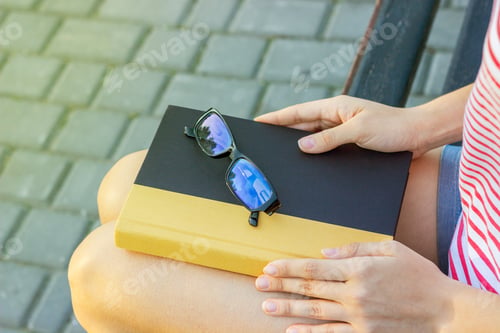 Preview: The girl on a bench in the park with a book and glasses in her lap.