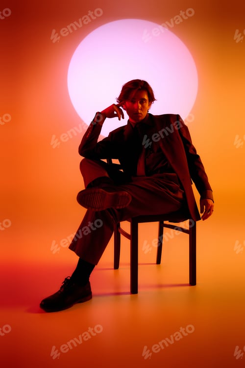 Preview: Stylish young man posed on a chair against a vibrant sunset backdrop in a studio setting