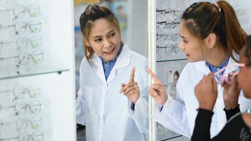 Preview: Smiling female optician helping child girl choosing a new eyeglasses frame in optical store.