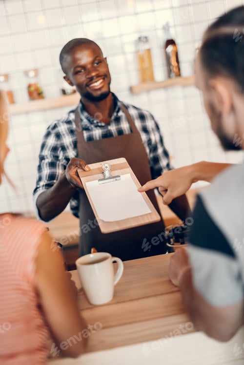 Preview: handsome smiling african american barista giving menu to clients on bar counter in cafe