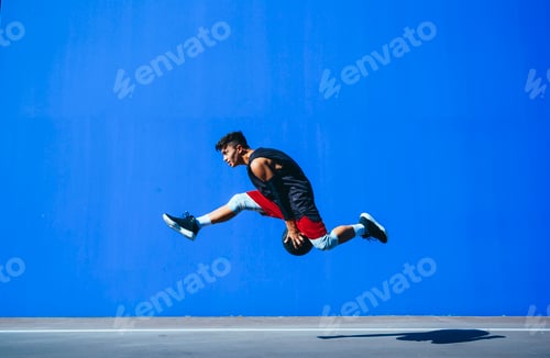 Preview: Side view of a young man holding a basket ball jumping in front of a blue wall.