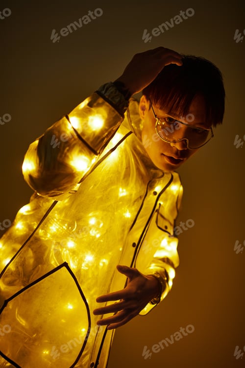 Preview: Stylish Asian man dressed in illuminated jacket poses confidently against warm backdrop