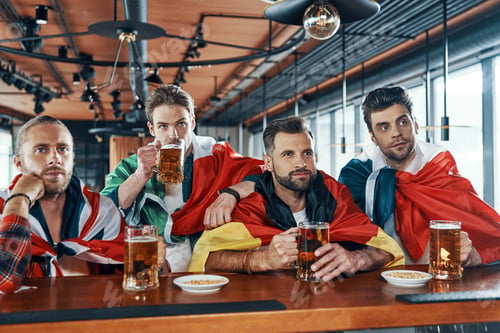 Preview: Young men covered in international flags drinking beer and watching sport game