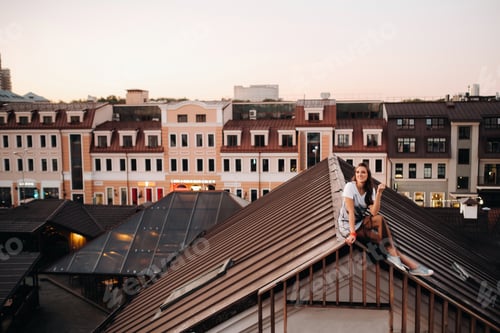 Preview: A girl sits on the roof of a house in the city in the evening. Portrait of a model in a dress and