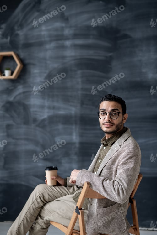 Preview: Contemporary businessman with glass of coffee sitting on chair in isolation