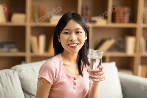 Preview: Happy asian lady holding glass of fresh clean water while sitting on couch in living room and