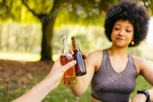 Preview: Two women cheering bottles of beer after workout in a green park