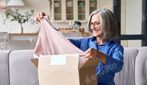 Preview: Senior mid age 60s woman opening parcel box at home on the couch.